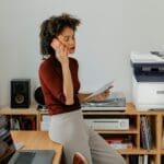 Impresora multifunción en color Xerox® C325 en un entorno de oficina en casa Una mujer sentada en un escritorio teniendo una llamada junto a una impresora Xerox® C325