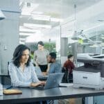 Impresora multifunción en color Xerox® C325 en una oficina Mujer profesional escribiendo en una computadora portátil en un escritorio en un entorno de oficina junto a una impresora Xerox® C325