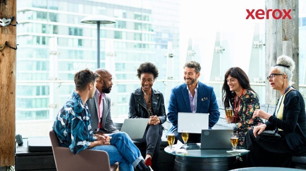 Un grupo de siete personas se sientan juntas, conversan y trabajan con sus computadoras portátiles con una ciudad como telón de fondo.