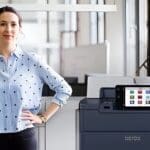 Una mujer de pie junto a la impresora Xerox® PrimeLink® Serie C9200 en un entorno de oficina moderno Una mujer de pie junto a la impresora Xerox® PrimeLink® Serie C9200 en un entorno de oficina moderno