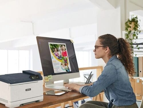 Una mujer sentada en un escritorio, concentrada en su trabajo, con una impresora Xerox® C320 a color