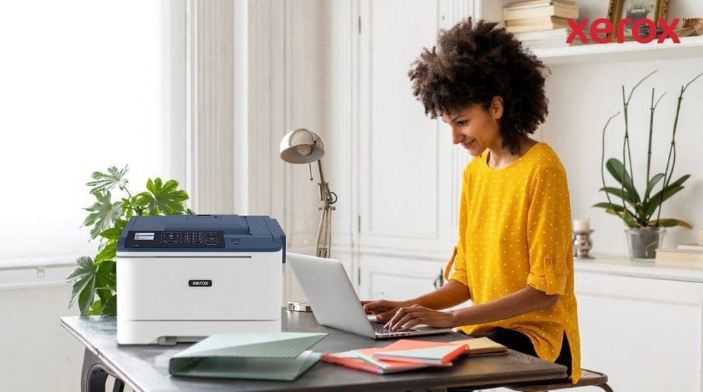 Una persona con cabello rizado sonríe mientras escribe en una computadora portátil en un escritorio, con una impresora compacta Xerox C310 ubicada al lado de la computadora portátil, mostrando una configuración de oficina hogareña moderna para la productividad.