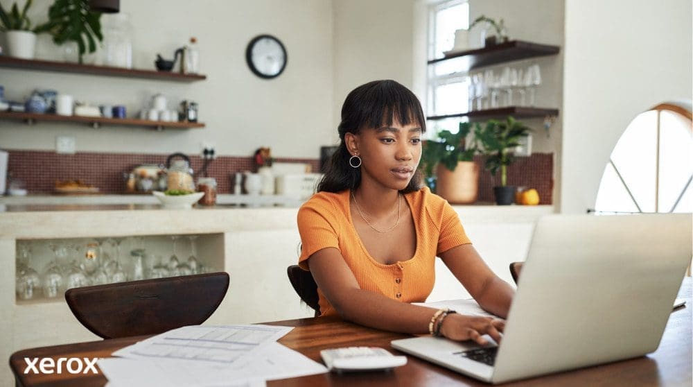 Xerox® DocuShare® Go: Impulsando el trabajo remoto Una persona está sentada en una mesa de cocina, concentrada en trabajar en una computadora portátil, con papeles y una calculadora cerca, lo que ilustra la flexibilidad de trabajar desde casa con una gestión eficiente de documentos.