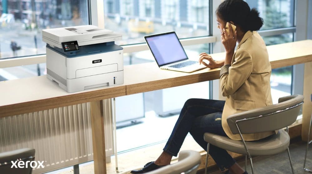 Experiencia de impresión y escaneo de Xerox®: cómoda y conectada Una persona está sentada en una mesa alta junto a un gran ventanal, hablando por teléfono mientras trabaja con una laptop. Una impresora multifunción Xerox B235DNI está convenientemente colocada sobre la mesa junto a ella, lo que sugiere facilidad de uso en un espacio de trabajo moderno.