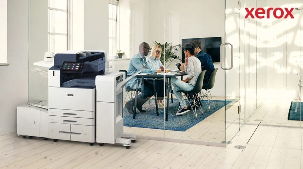 Una moderna impresora multifunción Xerox AltaLink B8245 se encuentra en primer plano en una oficina luminosa. Al fondo, un grupo de personas conversa en torno a una mesa.