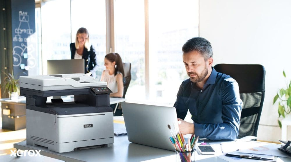 Xerox® Smart Start: diseñado para espacios de trabajo modernos Una persona está sentada en un escritorio, mirando una computadora portátil con una impresora multifunción Xerox C325 al lado. Al fondo, otras dos personas también trabajan en sus escritorios en un ambiente de oficina bien iluminado.