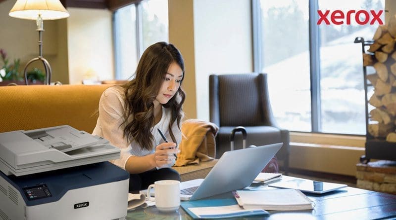 Una mujer trabaja con una computadora portátil en un cómodo entorno de oficina o sala de estar, con una impresora Xerox compacta visible en primer plano, lo que indica una integración perfecta de la impresión con los flujos de trabajo digitales.