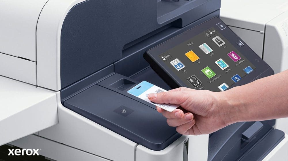 Xerox® Workplace Cloud y Suite: Autenticación segura e interfaz inteligente Una mano sostiene una tarjeta de seguridad y la escanea cerca del lector de tarjetas de una moderna impresora multifunción Xerox con una gran pantalla táctil que muestra varios íconos de aplicaciones.