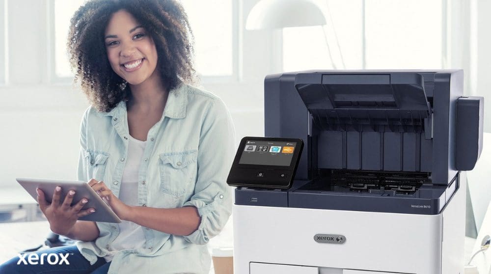 Xerox® Workplace Cloud y Suite: Integración fácil de usar Una persona sonriente está sentada junto a una impresora Xerox VersaLink B610 blanca, sosteniendo una tableta y mirando a la cámara, resaltando la naturaleza fácil de usar de las soluciones de Xerox.